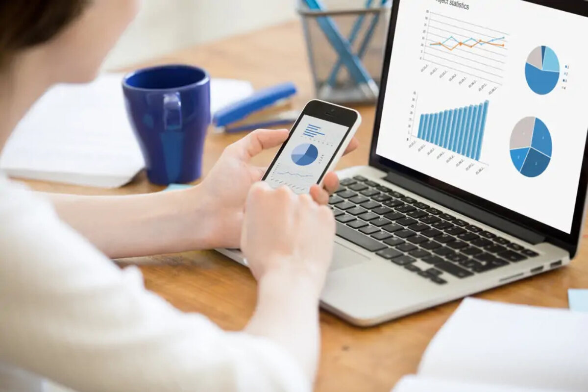 Businesswoman working on smartphone in front of open laptop, each screen showing graphs and charts