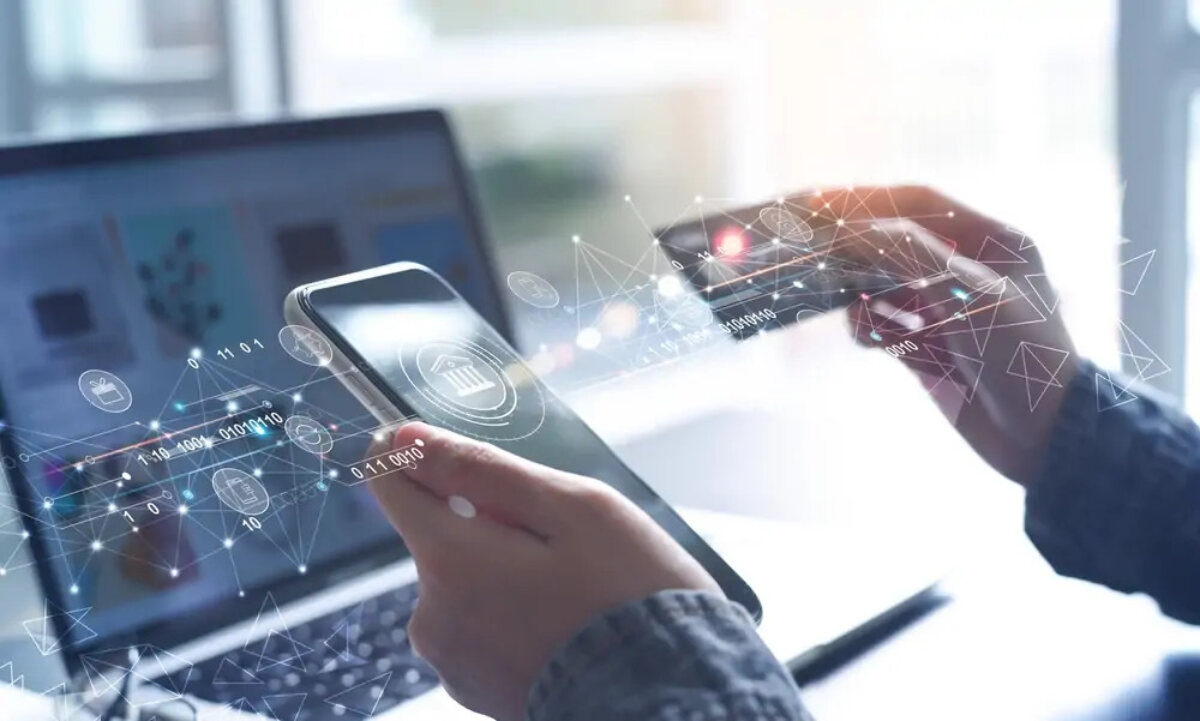 Businessperson using online banking app on smartphone and holding bank card above open laptop, overlaid with computer code images