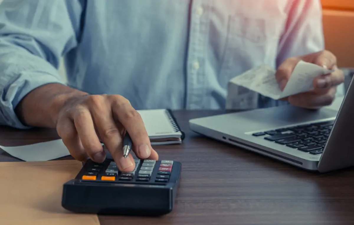 Businessman at desk in front of open laptop using calculator with one hand and holding receipt with the other to calculate VAT