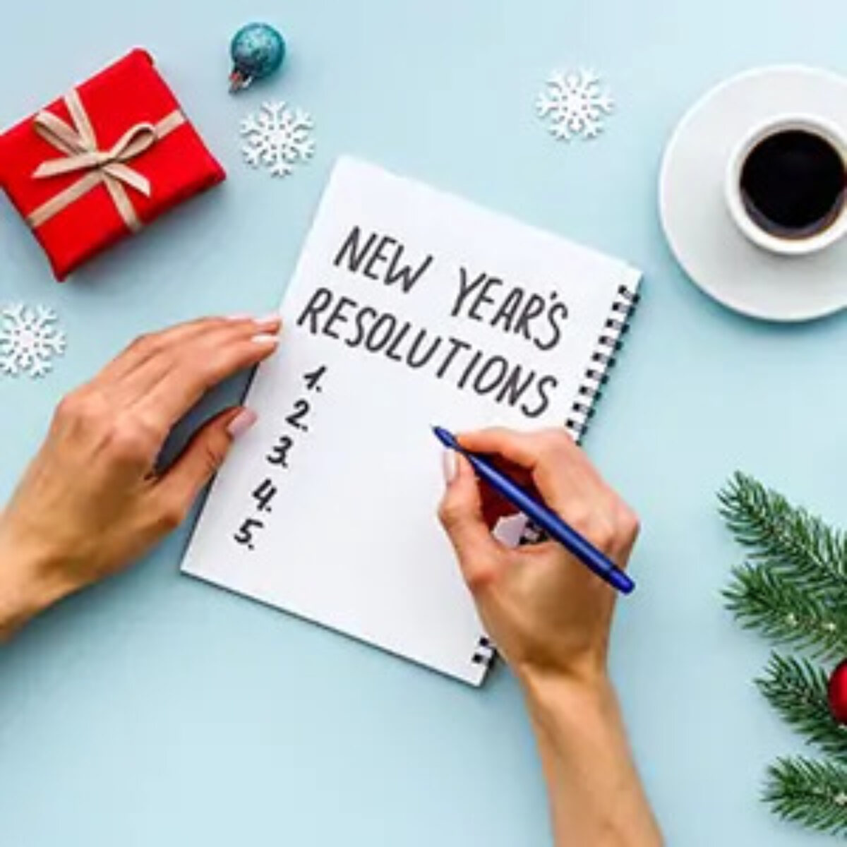 Hands writing list of New Year's resolutions in notebook surrounded by paper snowflakes, a miniature bauble, a miniature present, a fir branch and a cup of coffee