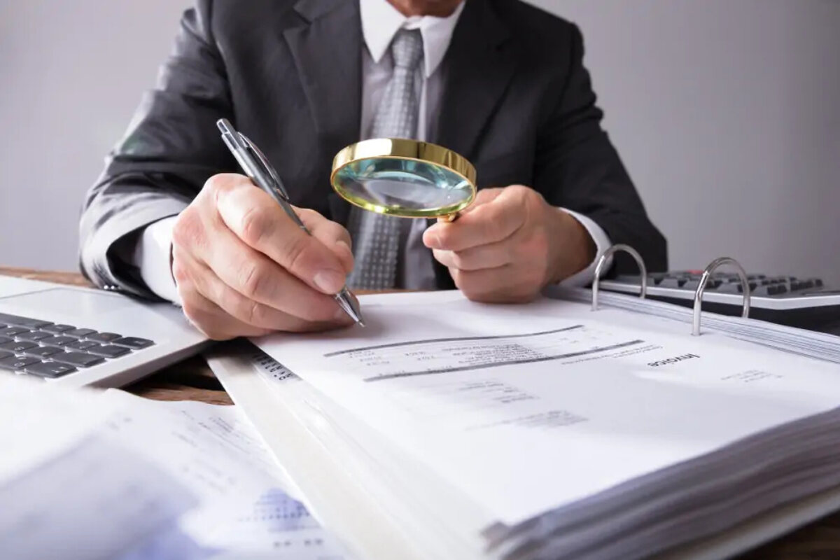 Businessman at desk holding pen and magnifying glass over invoice in ringbinder
