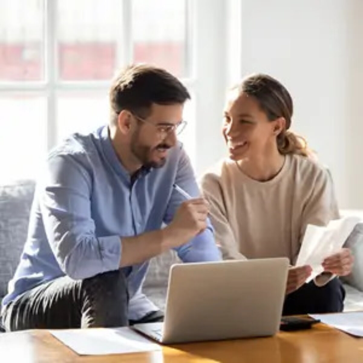 Two people sitting on sofa at coffee table with open laptop, holding documents and smiling