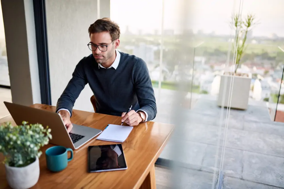 Businessman at desk holding pen over notebook and using laptop to calculate VAT