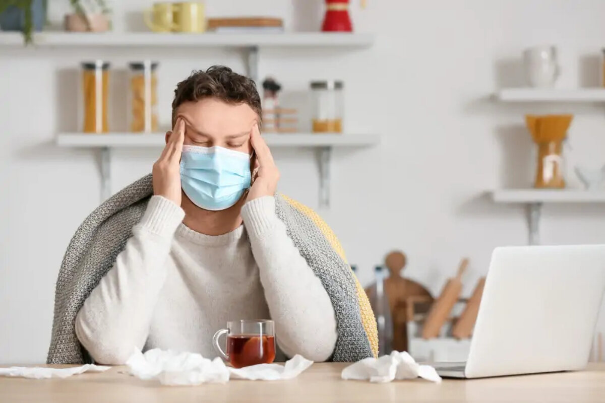 Ill man working from home on laptop wearing facemask, with eyes closed holding temples, surrounded by tissues and herbal tea