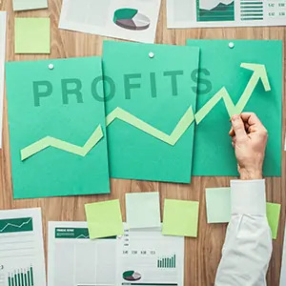 Businessman's hand holding an arrow showing increase over sheets of paper on a noticeboard with the word 'profits' across them