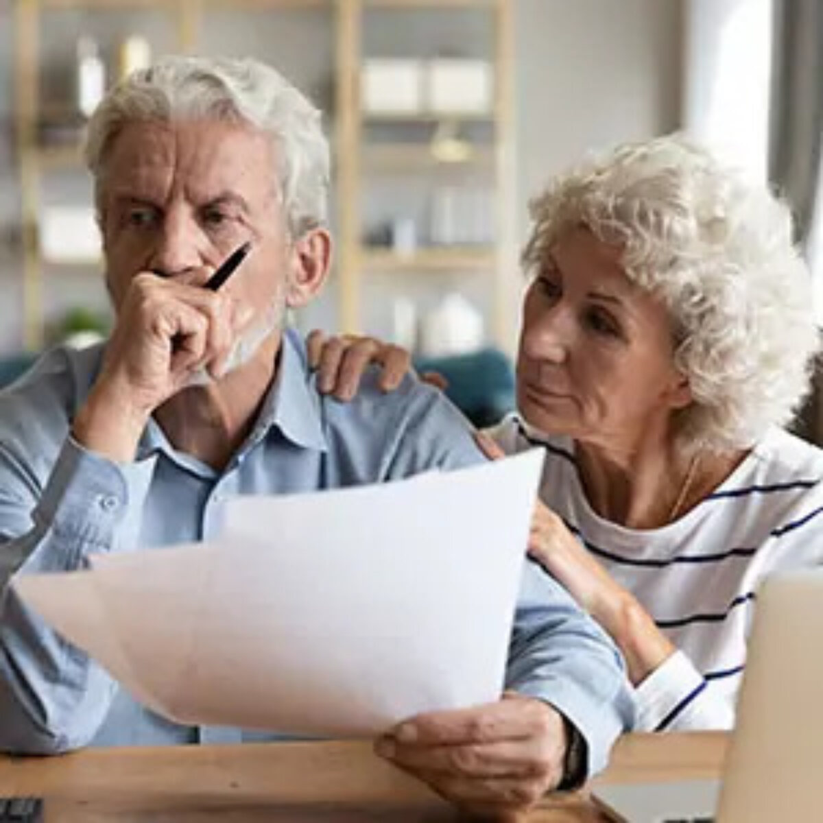 Elderly woman comforting elderly man who is holding pension document with fist to mouth looking into distance