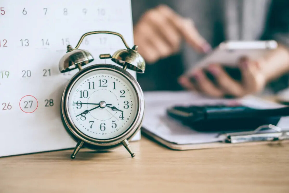 Alarm clock in front of calendar next to businesswoman using smartphone in front of clipboard and calculator