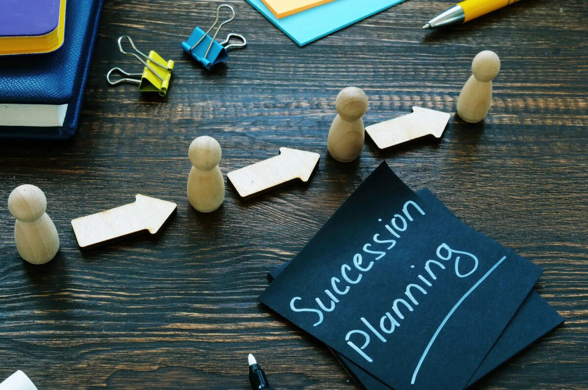 Wooden figurines in a row with arrows in between them next to a post-it note with 'succession planning' written on it