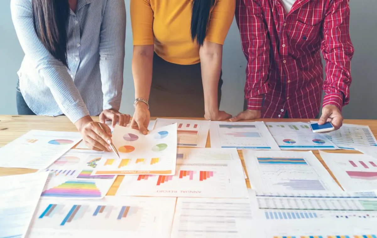 Three businesspeople standing over table looking and pointing at printed graphs and charts