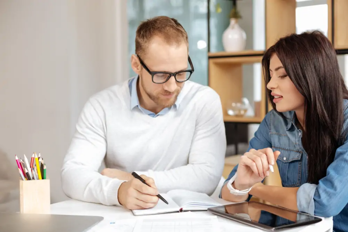 Two businesspeople sitting at table with tablet looking at open notebook