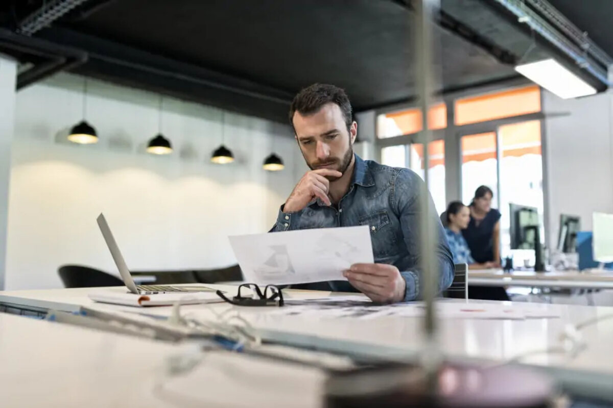 Man looking at document next to open laptop at desk in modern office