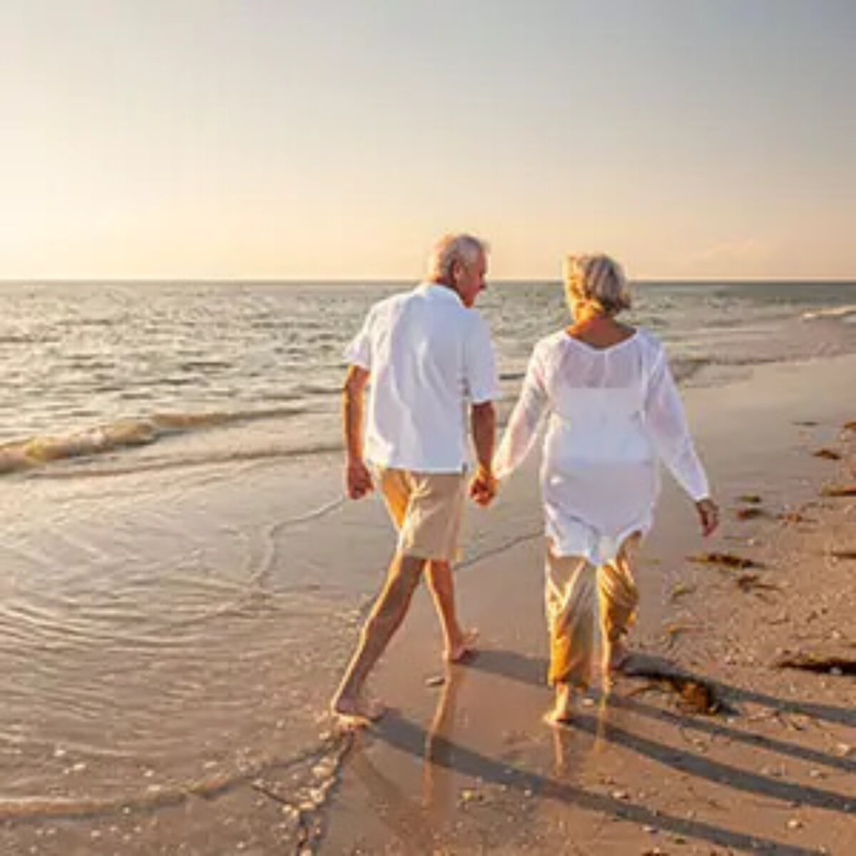 Elderly couple holding hands walking barefoot on sand next to sea