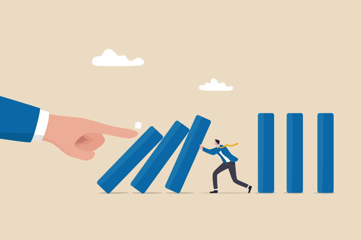 Cartoon of miniature businessman holding up toppling book spine-like rectangles that a giant finger has pushed over, with three more rectangles standing upright behind him