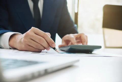 Businessman using calculator with one hand and holding fountain pen over document in the other making VAT calculations