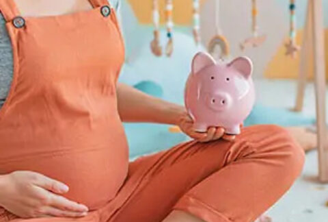 Pregnant woman sitting cross-legged on floor of nursery holding stomach with one hand and a piggy bank with the other