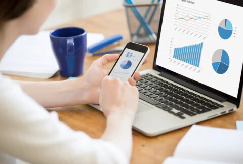 Businesswoman working on smartphone in front of open laptop, each screen showing graphs and charts