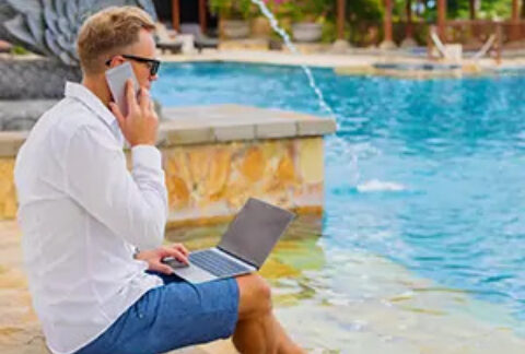 Businessman sitting on steps of outdoor swimming pool with laptop on lap and talking on phone