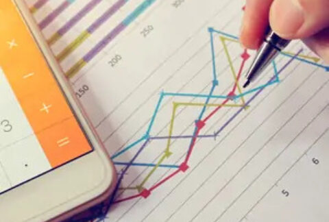 Hand plotting line graph on paper with pen and calculator app open on smartphone