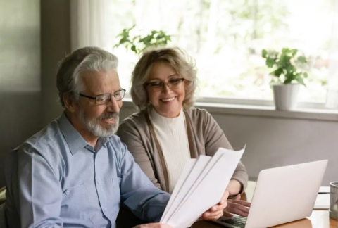 Elderly couple at table with open laptop holding and reading National Insurance and State Pension while smiling