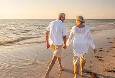 Elderly couple holding hands walking barefoot on sand next to sea