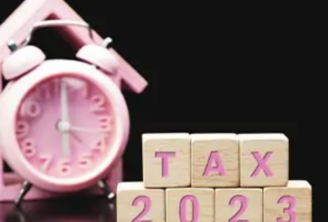 Wooden blocks with letters on them arranged to spell out 'tax 2023' in front of wooden house silhouette and alarm clock
