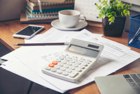 Calculator on desk on top of documents next to pencil, smartphone and cup of coffee