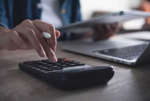 Businessperson calculating tax at desk while holding tablet ad using calculator next to open laptop