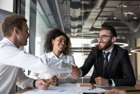 Two businessmen shaking hands over table at meeting and one businesswoman sitting between them