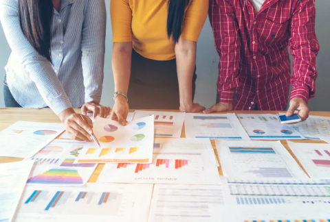 Three businesspeople standing over table looking and pointing at printed graphs and charts