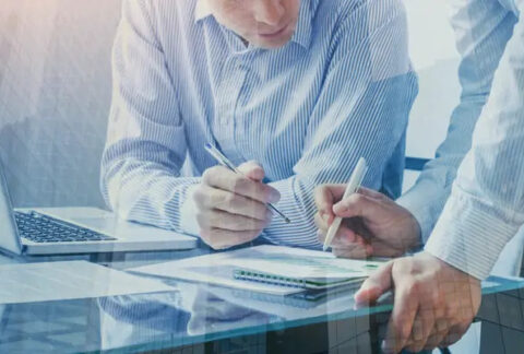 Two businessmen at desk with open laptop looking at and writing on Corporation Tax document