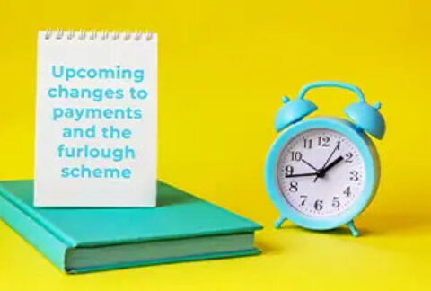 Alarm clock next to closed book with open notebook on top with page displaying 'upcoming changes to payments and the furlough scheme'