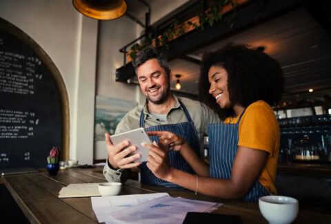 Two baristas behind a coffee bar holding and looking at a tablet
