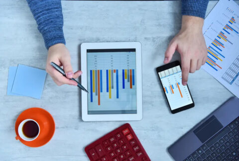 Businessman holding pen to tablet showing bar chart with one hand and using smartphone displaying similar bar chart with the other