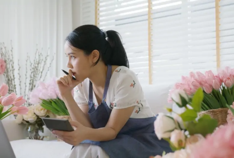 Florist sitting in front of laptop in shop with calculator in hand making cash flow calculations