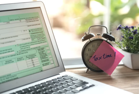 Alarm clock with post-it note on face saying 'tax time!' next to laptop with screen displaying Self Assessment form