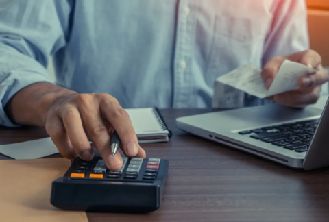 Businessman at desk in front of open laptop using calculator with one hand and holding receipt with the other to calculate VAT