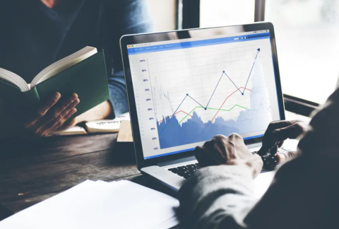 Businessperson at table using laptop with business growth graph displayed on screen opposite another businessperson reading a book