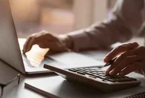 Businessperson at desk using laptop with one hand and calculator with other