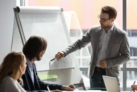Businessman standing pointing at flip chart with pen and talking to colleagues sitting down in business finances meeting