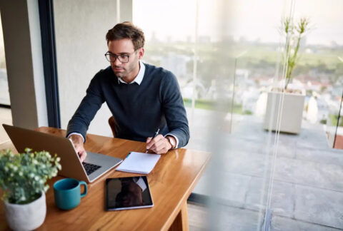 Businessman working on laptop and notebook and pen at desk next to tablet