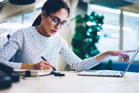 Businesswoman at desk with pen in one hand and pointing at laptop screen with the other looking at management accounts