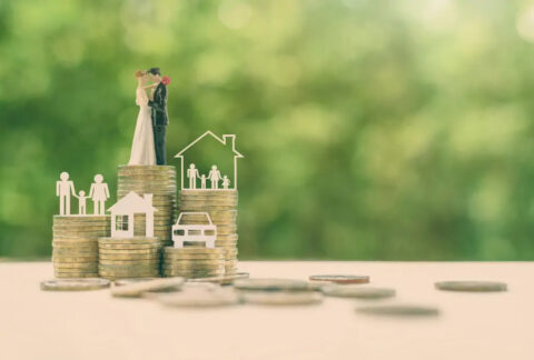 Five stacks of coins of varying heights, one with a paper cutout of a car on top, one a house, one two parents and a child, one a house with a family inside, and one a wedding cake topper