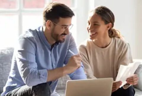 Two people sitting on sofa at coffee table with open laptop, holding documents and smiling