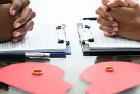 A separating couple opposite each other with their hands clasped over separate clipboards and a broken paper heart in front with wedding rings on top
