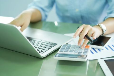 Businessperson at desk using calculator with one hand and laptop with the other to make payroll calculations