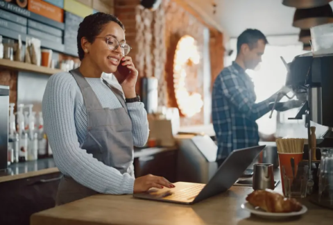 Cafe owner taking phone call and using laptop behind coffee bar