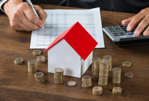 Businessman doing calculations beside a model house and stacks of coins