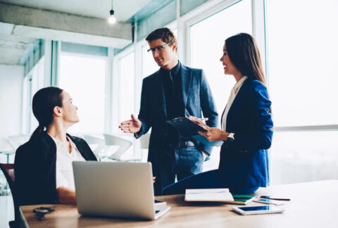 Businesswoman sitting at desk talking to two colleagues standing next to her