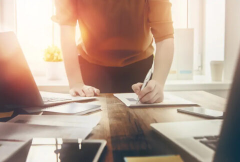 Business woman standing while writing on a document on desk next to laptop
