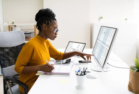 Businesswoman at desk pointing at computer screen and writing on document in ring binder